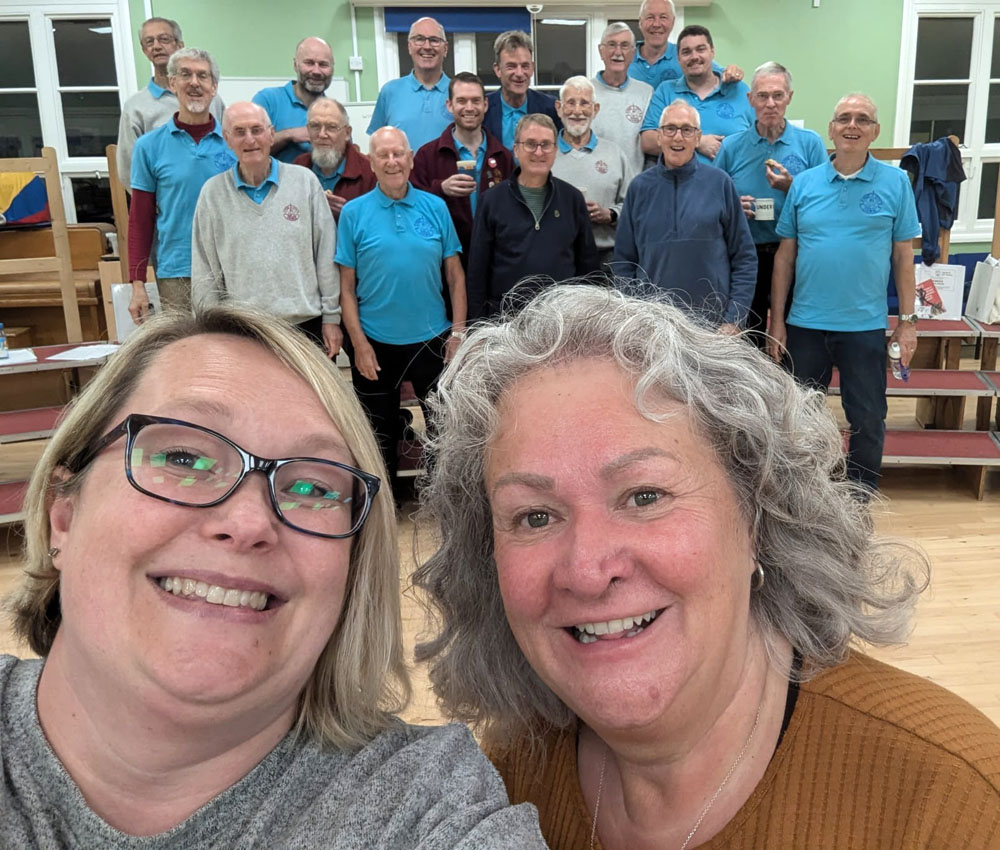 Shanna from BABS and Carol of FCC take a selfie, with the chorus members forming a glamorous backdrop